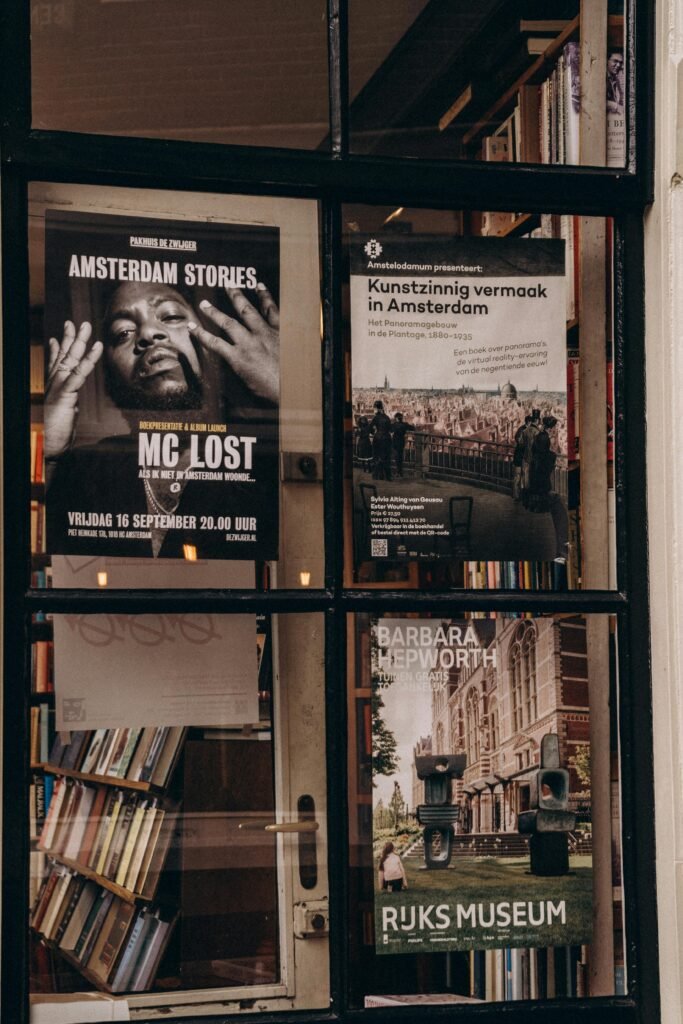 Posters and books seen through a bookstore window in Amsterdam featuring art and event advertisements.